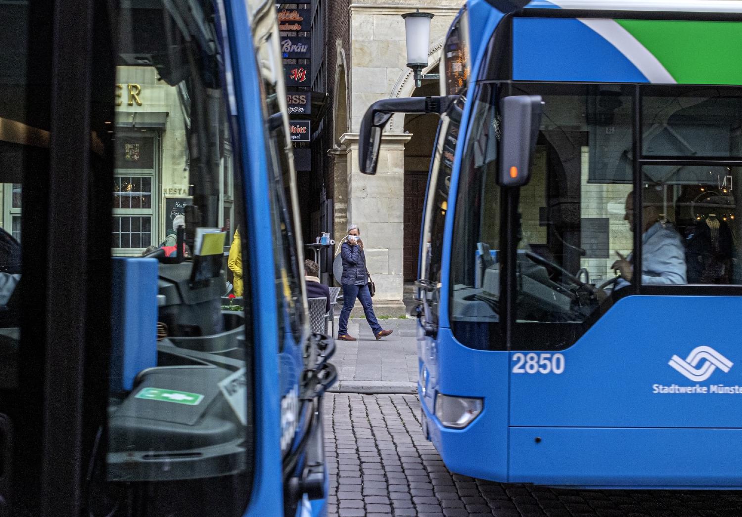 Blick zwischen zwei Bussen hindurch, Frau hält sich eine OP-Maske notdürftig vor Mund und Nase