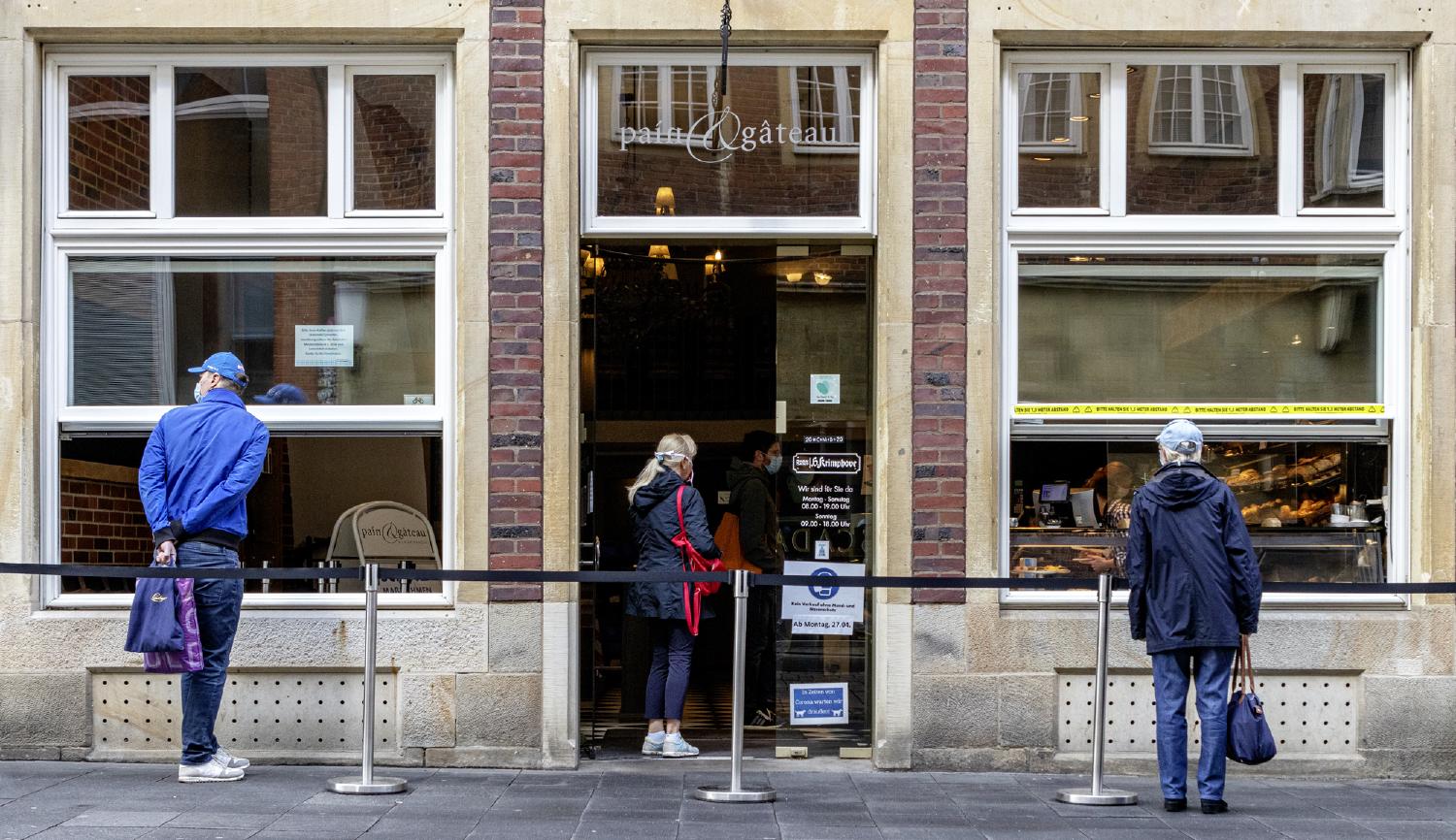 Einelne, der Kamera abgewandte, Personen warten mit viel Abstand im Außenbereich einer Bäckerei
